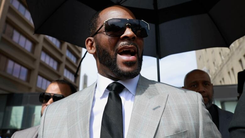 A man in a suit and large black sunglasses walks through a downtown area underneath an umbrella. 