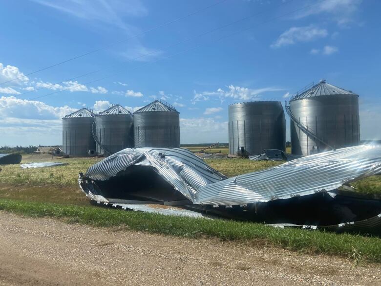 In the foreground, lying next to the edge of a gravel road, is the battered husk of a metal grain bin. Lying on its side, the metal is dented and stretched out as though the walls were unwrapped from their moorings. In the background sit a row of five other bins, with a gap between the third and fourth where the damaged one may have previously stood. Damage is apparent on the background bins; one is missing a roof completely, while others appear to have had their winding staircases torn off. Metal debris is scattered around the midground of the photo.