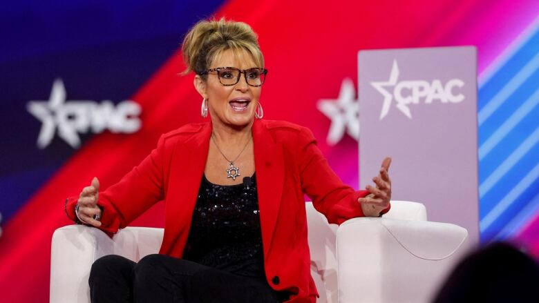 A woman in a red blazer speaks and gestures while sitting on an armchair on a stage.