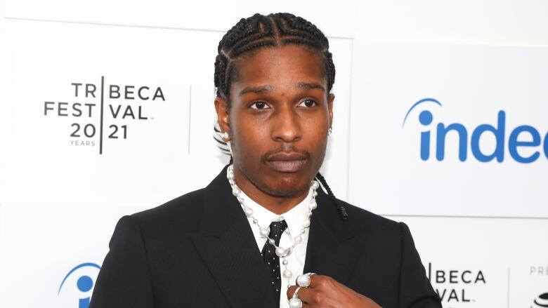 A man wearing a black suit and marble necklace stands in front of a backdrop with the words 