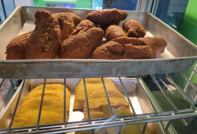 A metal wrack is filled with two trays of food: One is filled with half-moon-shaped, yellow hand pies; the other has oblong fried bread.