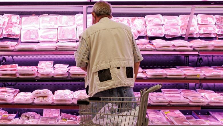 A man stands at a supermarket meat section with his back to the camera, looking at the packaged meat. 