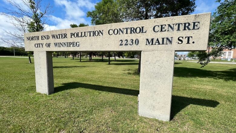 A stone sign stands on green grass in front of trees that says 