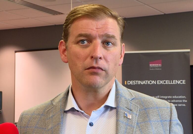 A head and shoulders shot of a person in a light grey blazer. Behind them a sign with a Memorial University logo can be seen.