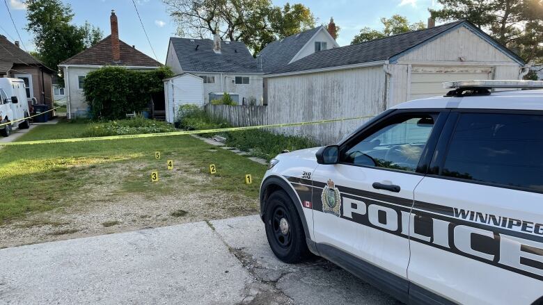 Five evidence markers can be seen in the backyard of a small bungalow, which has police tape around it and a police cruiser parked in the lane near a garage on the right side of the photo.