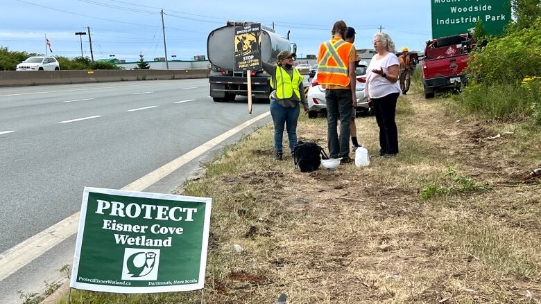 People stand on the side of a highway near a protest sign.