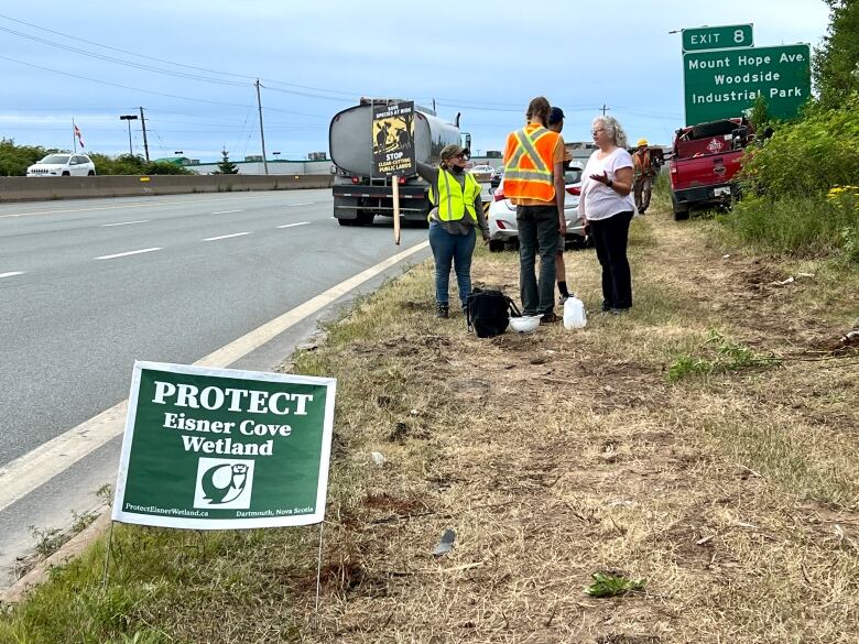 People stand on the side of a highway near a protest sign.