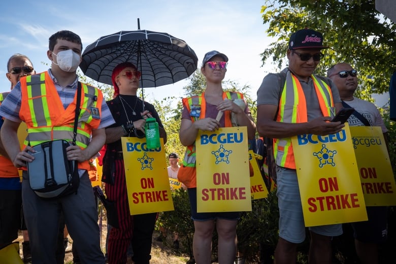 A number of people at a picket line holding yellow signs that read 'BCGEU On Strike'.