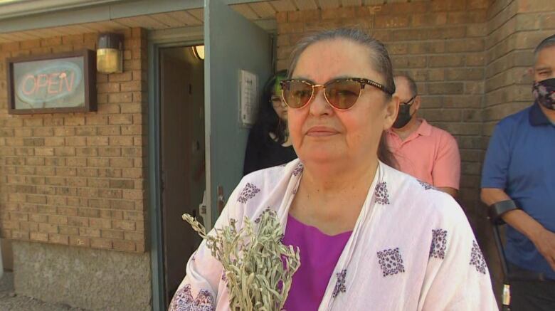 A woman wearing sunglasses, a purple shirt and a white and black shawl holds a bundle of sage in front of a brick building.