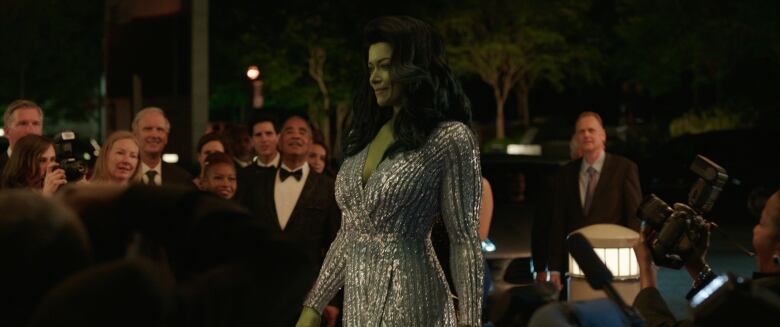 A tall, green-skinned woman in a glittery dress strides past a crowd of onlookers. 