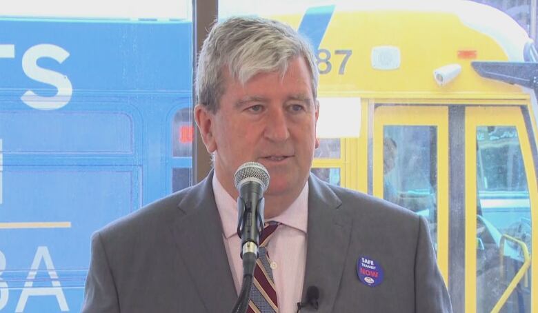 A man in a suit jacket and tie speaks into a microphone. Behind him, a Winnipeg Transit bus drives by.