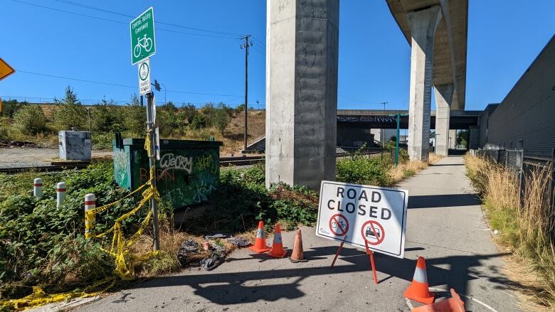 A 'Road Closed' sign and orange traffic cones litter a bike path next to train tracks.