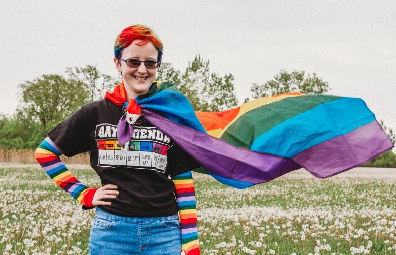 Woman stands in a field of dandelions wearing a Pride flag as a cape.