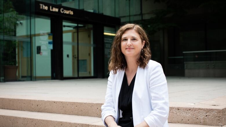 A woman wearing a white coat sits on steps outside a courthouse.