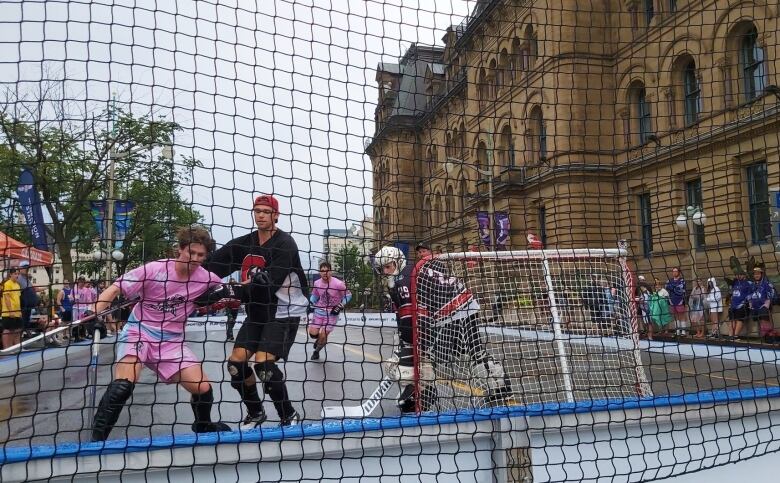 Two street hockey players fight for a ball behind a mesh screen on an outdoor street while a goalie looks on.