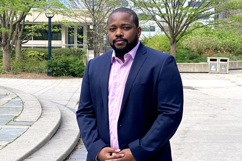 A Black man in a suit jacket stands in a city park. 