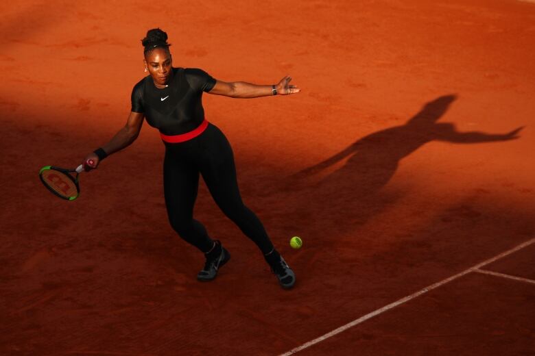 A woman standing on a tennis court, seen from above, begins a swing at a tennis ball.