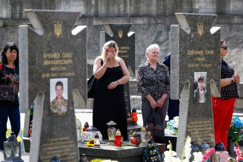 A woman wipes tears alongside others in front of large monuments in a cemetery.