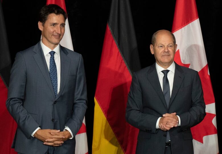 Two standing men wearing business suits clasp their hands in front of the German and Canadian flags 