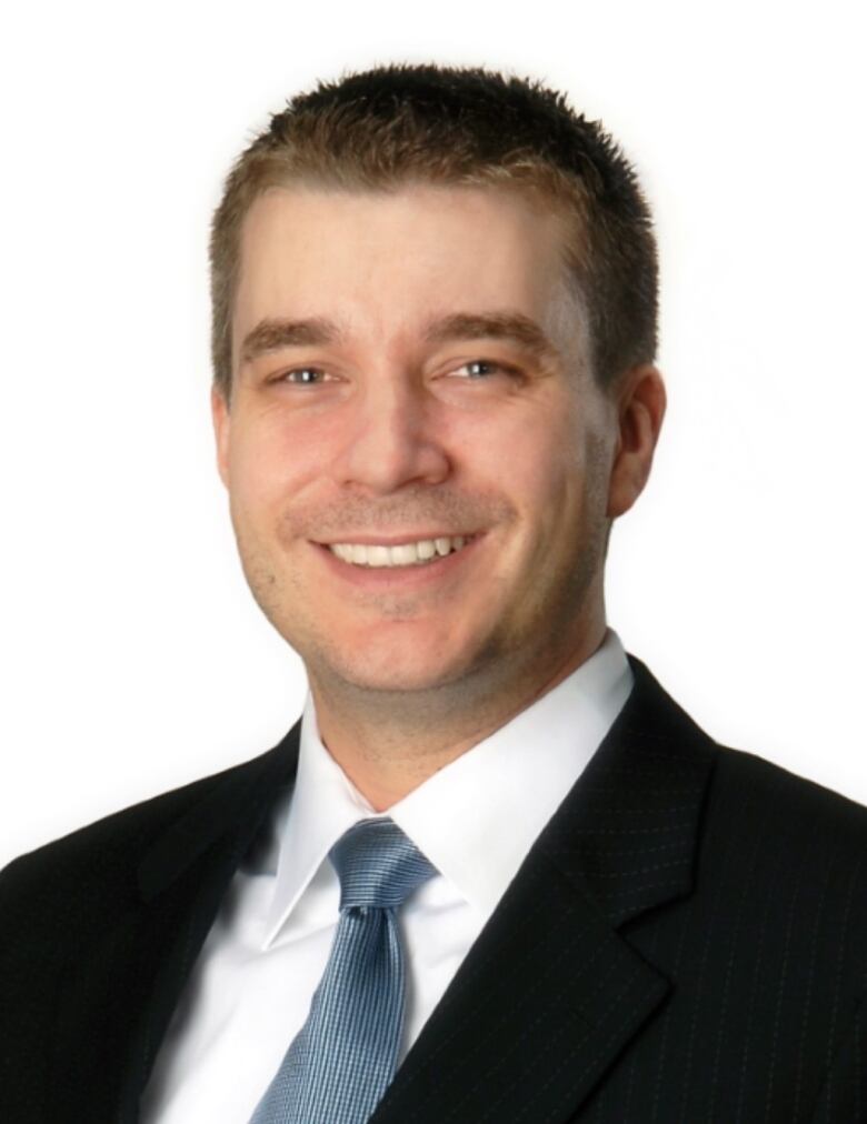 A head shot of professor Nicholas Carleton. He is wearing a black suit, white shirt, and patterned light blue tie. He stands before a white background.