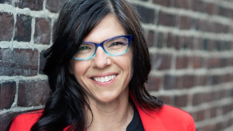 A shot of a smiling person with long dark hair, wearing glasses and a red blazer. 