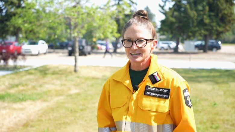 A woman wearing a yellow uniform stands outdoors, with parked cars and grass in the background.