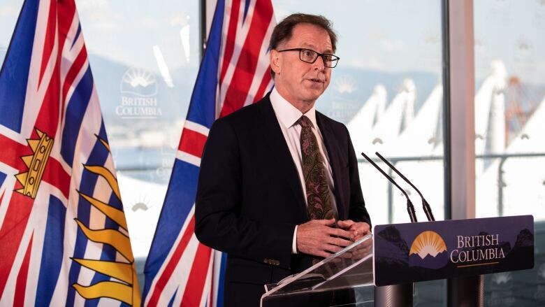 Adrian Dix, a white man with a black tie and a black coat on, talks at a podium marked 'British Columbia'. The flags of B.C. and the U.K. are behind him.