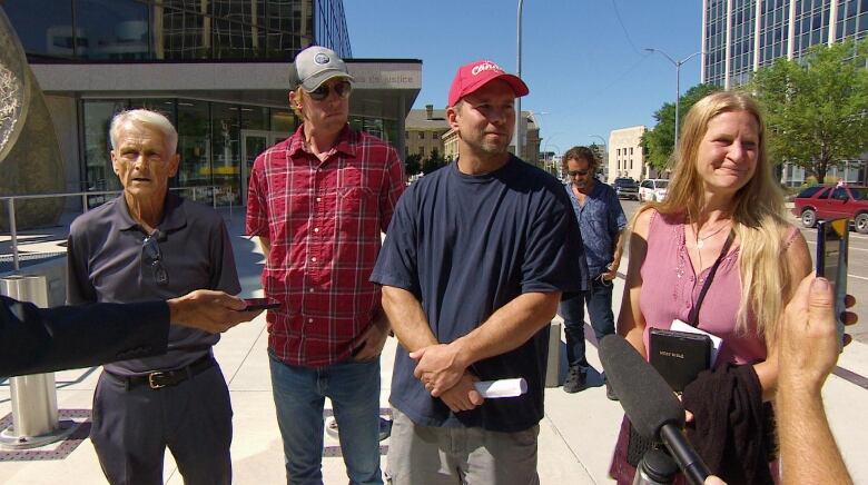 Four people, three men and one woman, stand in front of a large office building with hands holding microphones and cellphones in the foreground.