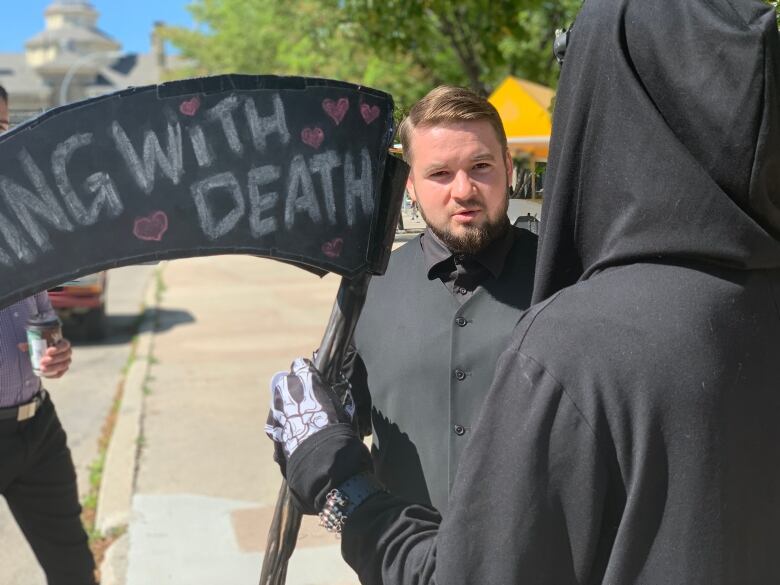 A man with a beard and wearing black clothes speaks to a person dressed as the Grim Reaper, holding a plastic scythe with the letters 