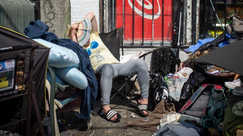 A person is sitting on a foldable chair among tents and debris on a pavement. They are carrying a hand fan over their face.