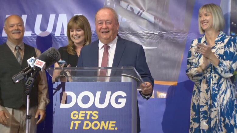 Doug McCallum, a bald white man wearing a red tie, smiles at a podium that reads 'DOUG GETS IT DONE'. He is flanked by smiling supporters.