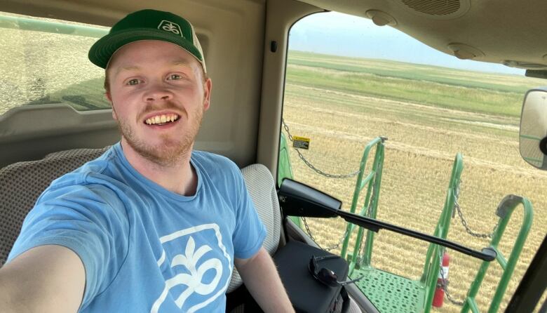 A man wearing a blue shirt  and green baseball bat takes a photo in the seat of a combine.