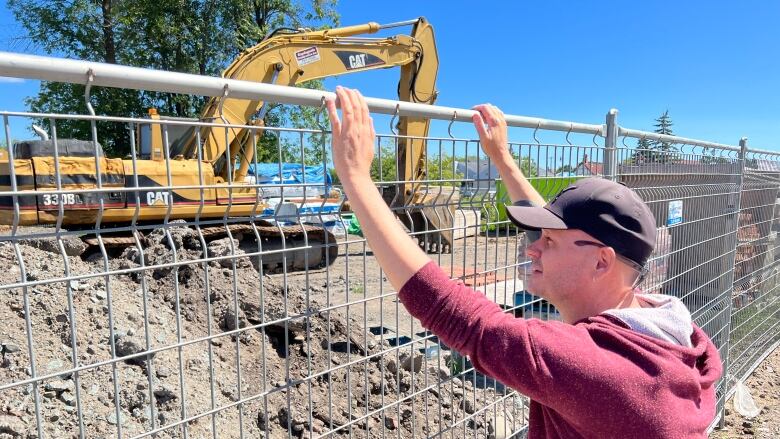 A man leans up against a fence and watches a machine dig at a construction site. 