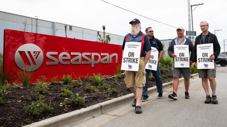 A group of men with 'ON STRIKE' placards hanging on their chests walk past a sign that reads 'Seaspan'.