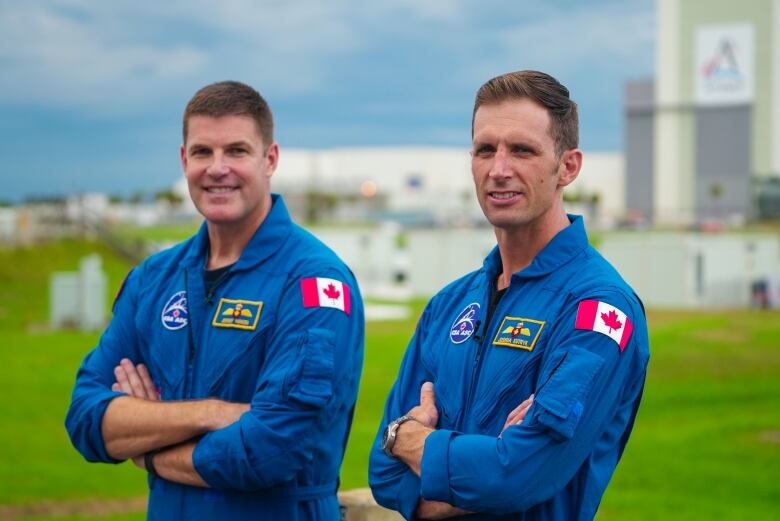 Canadian astronaut Jeremy Hansen, left, and Joshua Kutryk, right, stand in their blue flight suits, with bright green grass in the background along with several NASA buildings.