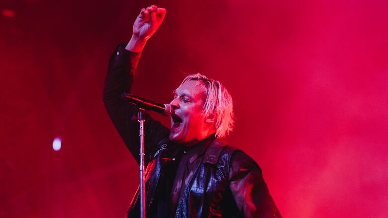 A man in a leather jacket holds an electric guitar and sings into a microphone, against a red light background.