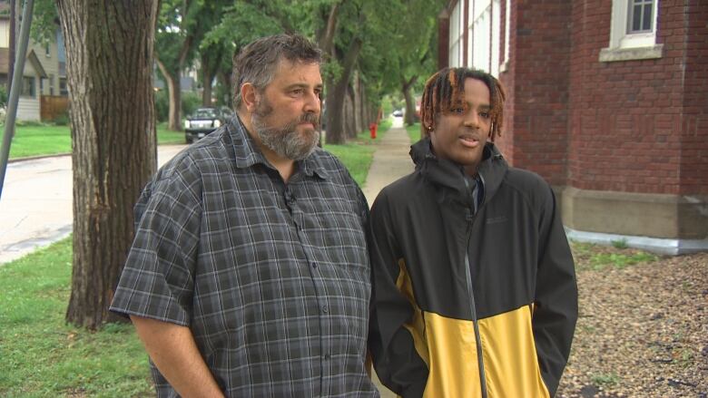 A man and a teenage boy stand next to each other, with serious looks on their faces. The man is speaking, while the teen has his hands in his pockets.