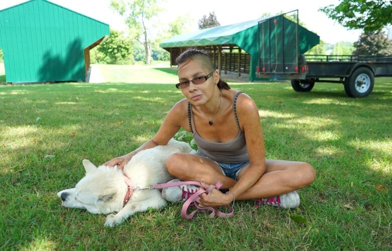 A woman sits in a grassy field with her white dog at her side.