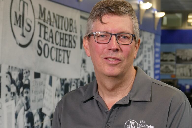 A man with glasses wears a polo shirt that says 'Manitoba Teachers' Society.' He stands in front of a banner that says the same thing. It features black-and-white photos of people gathered together and holding protest signs about education.