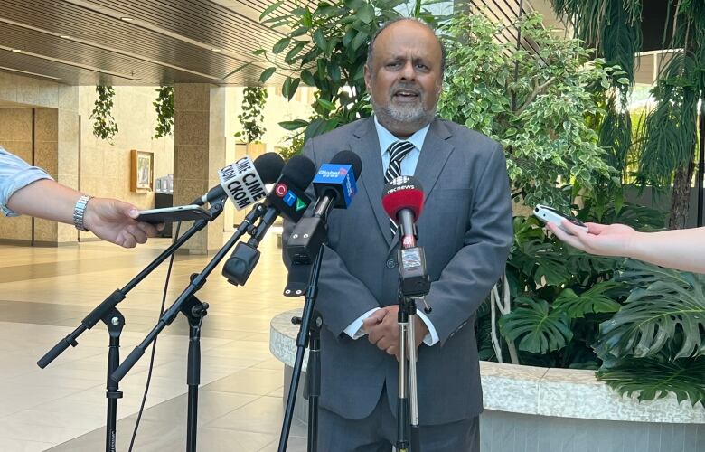 A man in a grey suit speaks in front of several news media microphones.