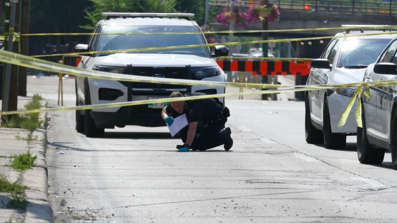 A Winnipeg police investigator wearing latex gloves and holding a piece of paper kneels as he examines something on the street, behind yellow police tape and surrounded by three police cruisers.