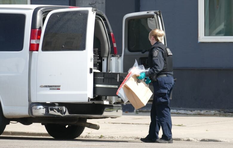 Winnipeg police officer holding a large paper bag stands facing the back of a white police van with its back doors open.