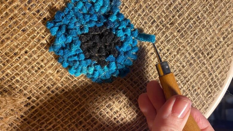 A bright blue flower is shown on burlap with a hand holding a hook.