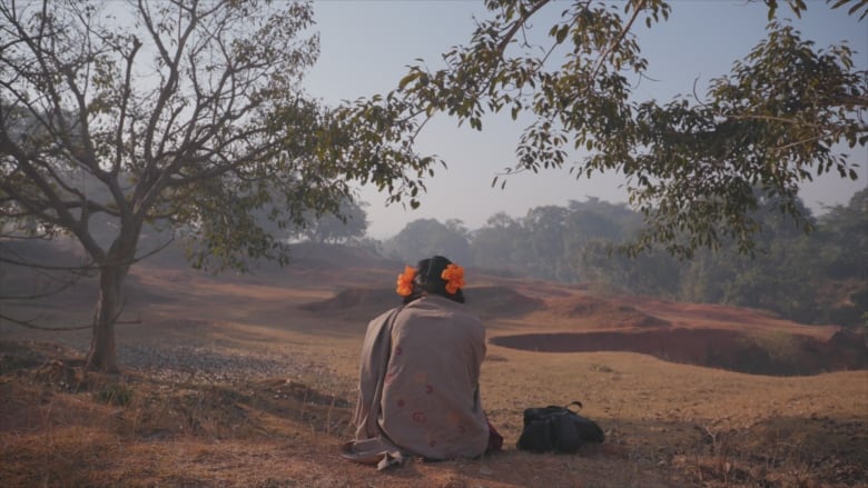 A girl with bright hair ties sits facing away from the camera. She is sitting among trees and barren ground. 