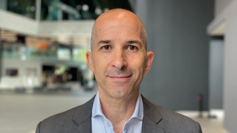 A man with no hair and a suit stares at the camera with a blurred background.