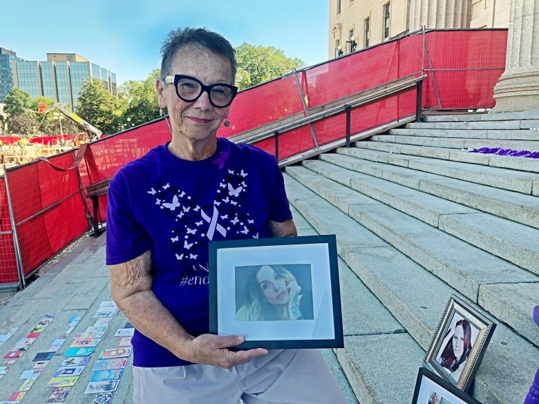 A woman wearing a purple shirt holds a picture frame containing a photograph of her daughter. Other picture frames sit on steps leading to a large stone building.