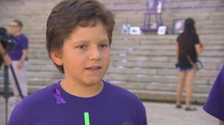 A young boy with brown hair wears a purple shirt and a purple ribbon.