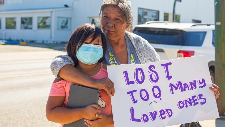 An older woman wraps her arms around a child while holding a sign that says 