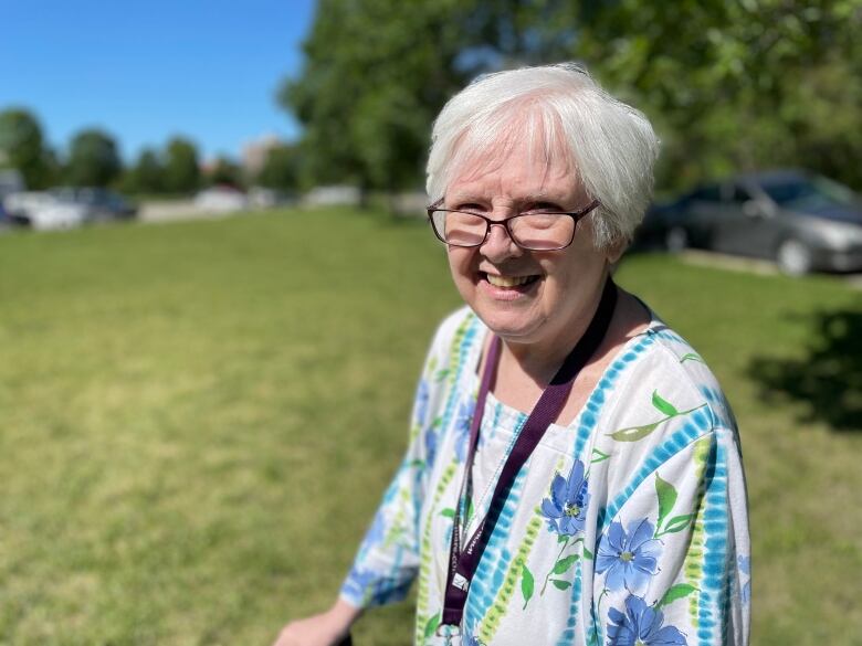 A woman with short white hair and glasses smiles while standing in a green space.