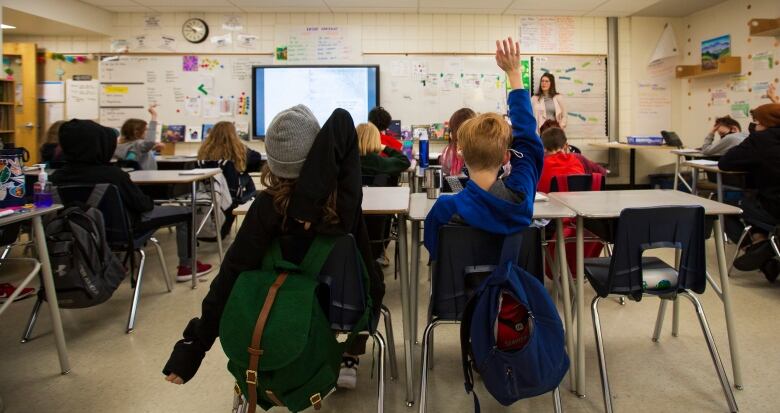 Students are seen interacting with their teacher and putting their hands up in a brightly lit classroom.